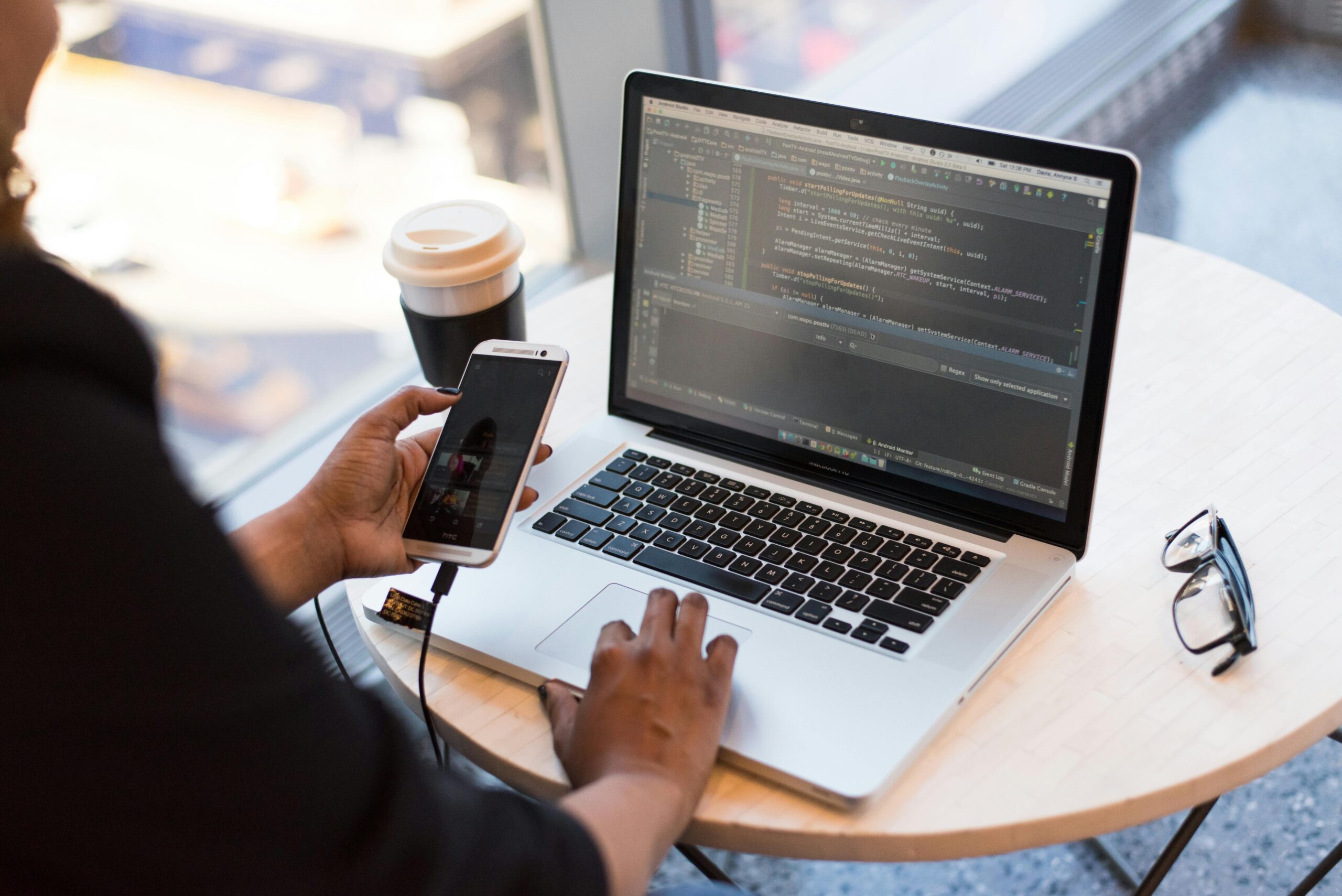 A person holding a phone and writing code for developing an app on a laptop.